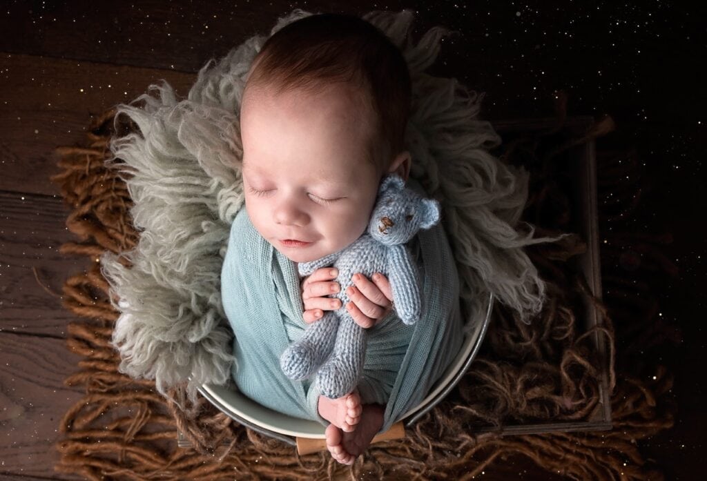 Older newborn cuddling teddy in bucket with blue wrap