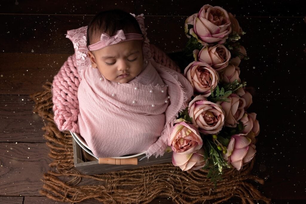 older newborn wrapped in pink wrap sitting in a bucket with pink flowers